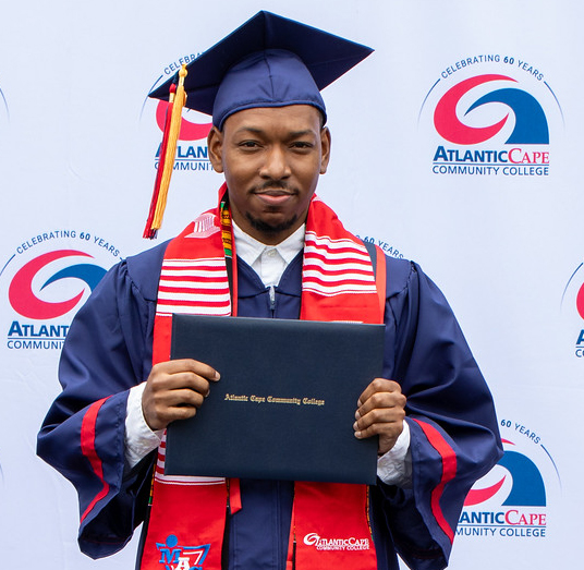 Navin, a Men of Atlantic Cape (MAC) graduate, wears his cap and gown while holding his diploma at graduation.