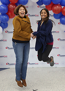 two people jumping up while holding hands in front of a backdrop with red, white, and blue balloons. 